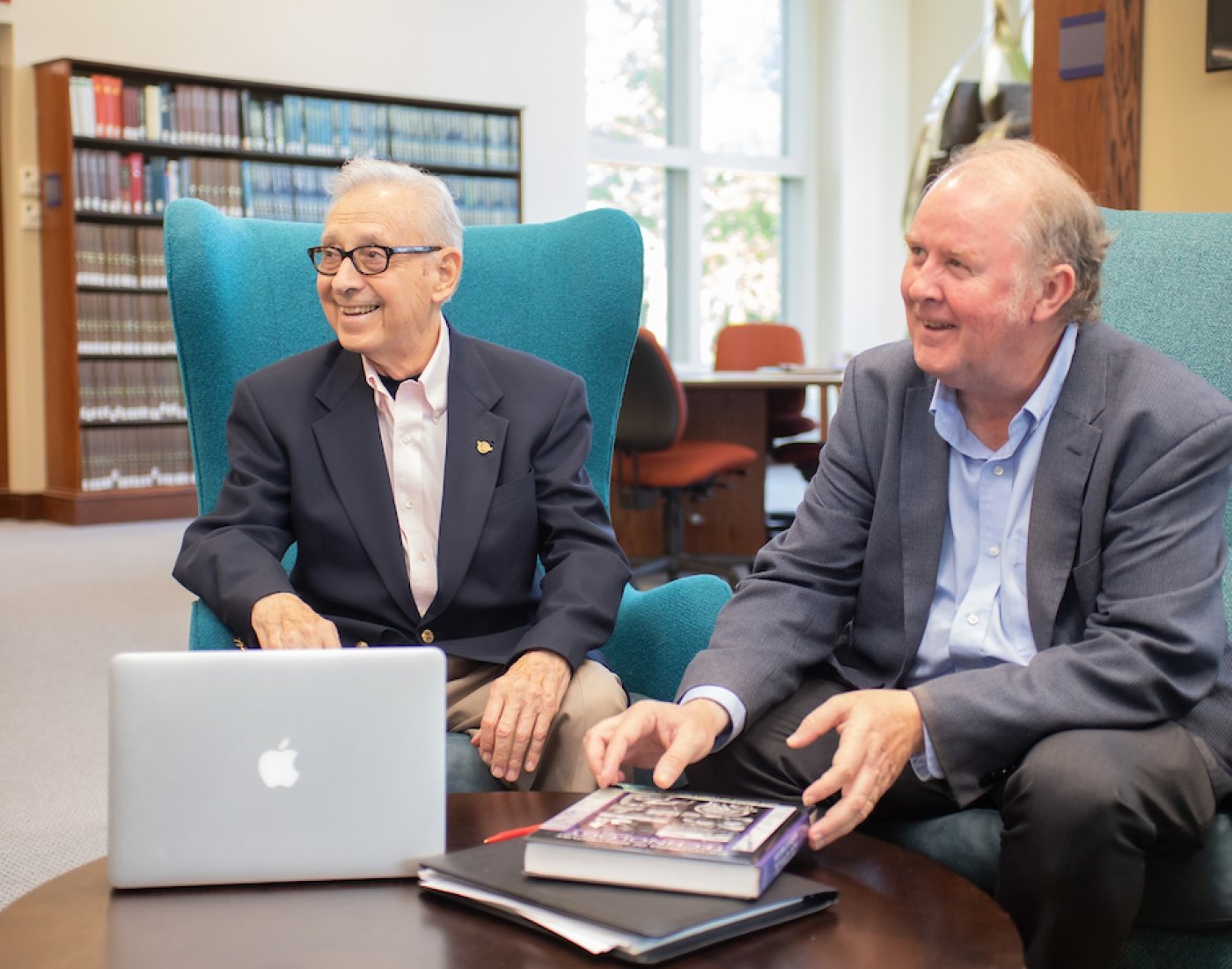 Two older gentlemen smiling in a library with laptops and books