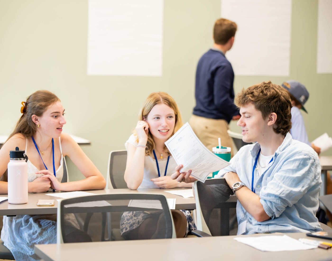Male student is holding his work paper and sharing with two female students while another student is in background adding comments to paper on wall