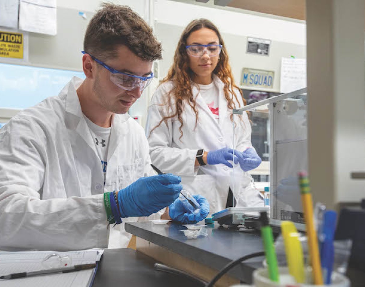 Male and Female student working in lab, wearing white coats and gloves handling test tubes