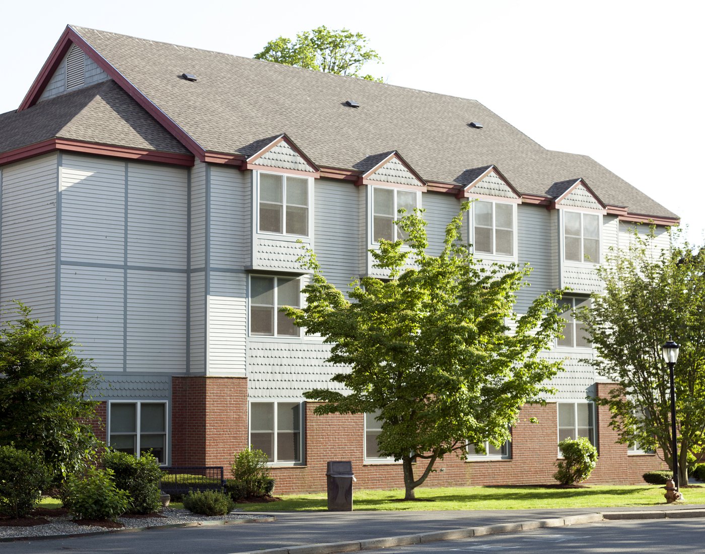 Three-story residential building with light siding, brick lower walls and trees in front.