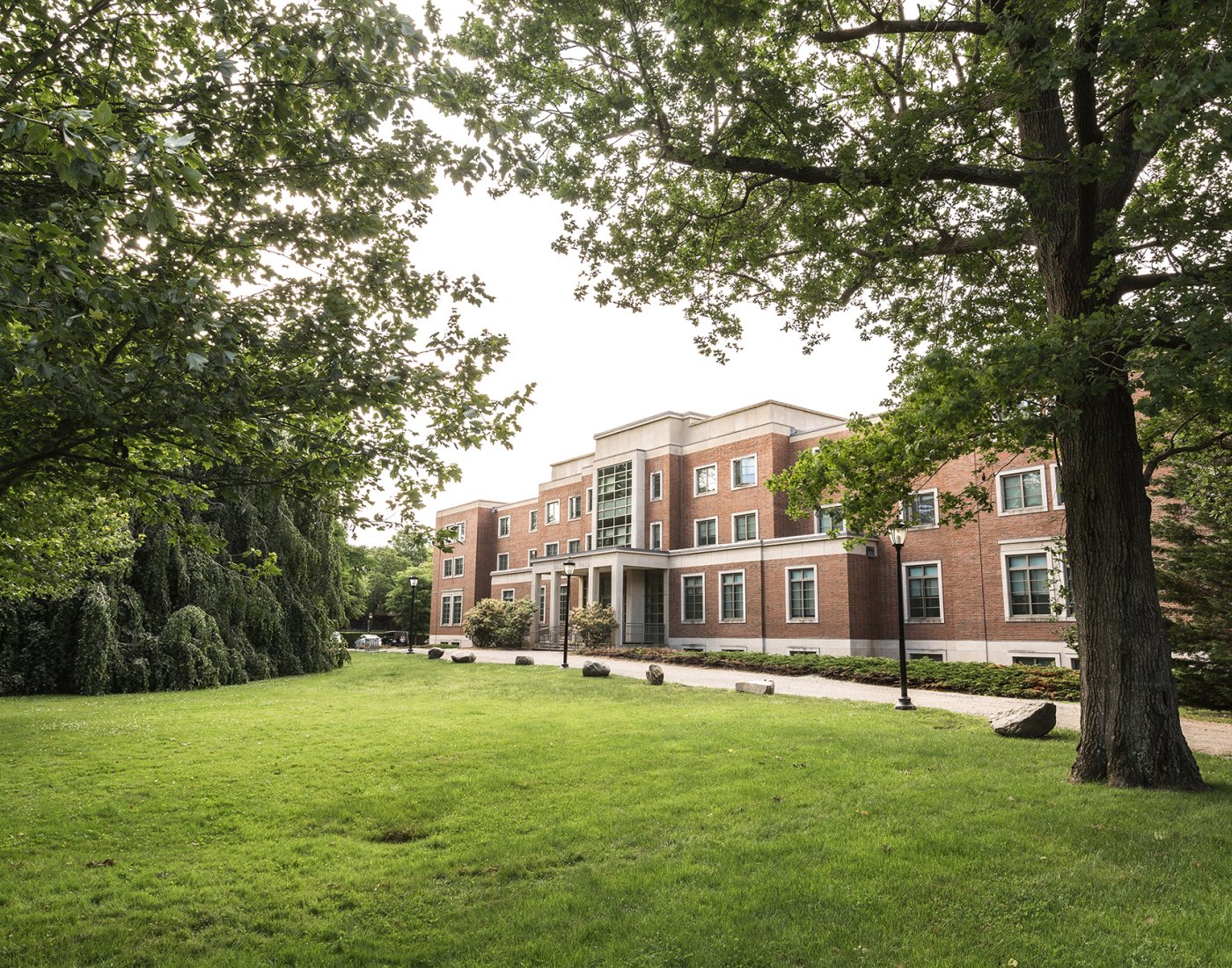 A brick building with large windows and a columned entrance, surrounded by a green lawn and tall leafy trees.