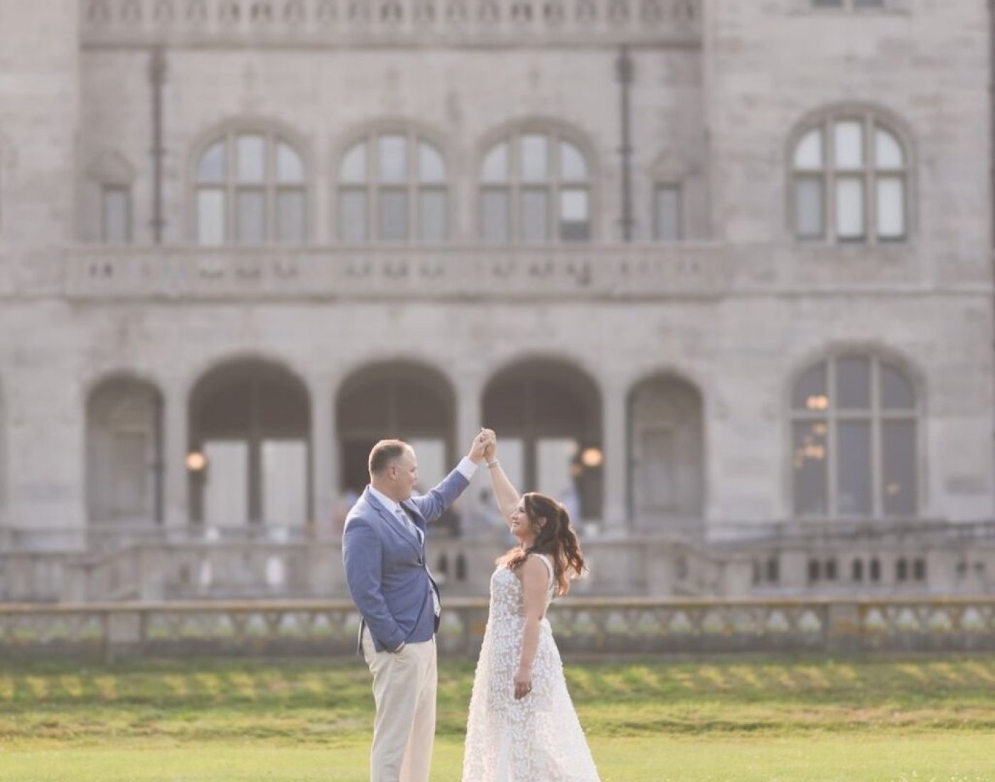 A couple in wedding attire stands on a lawn in front of a large historic stone mansion.