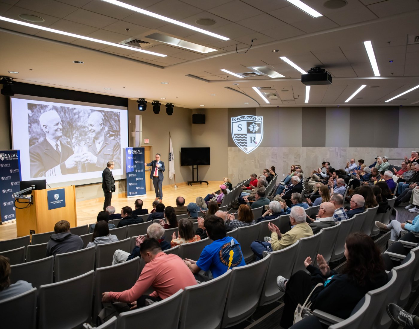 An auditorium full of people looking at a screen with political figures