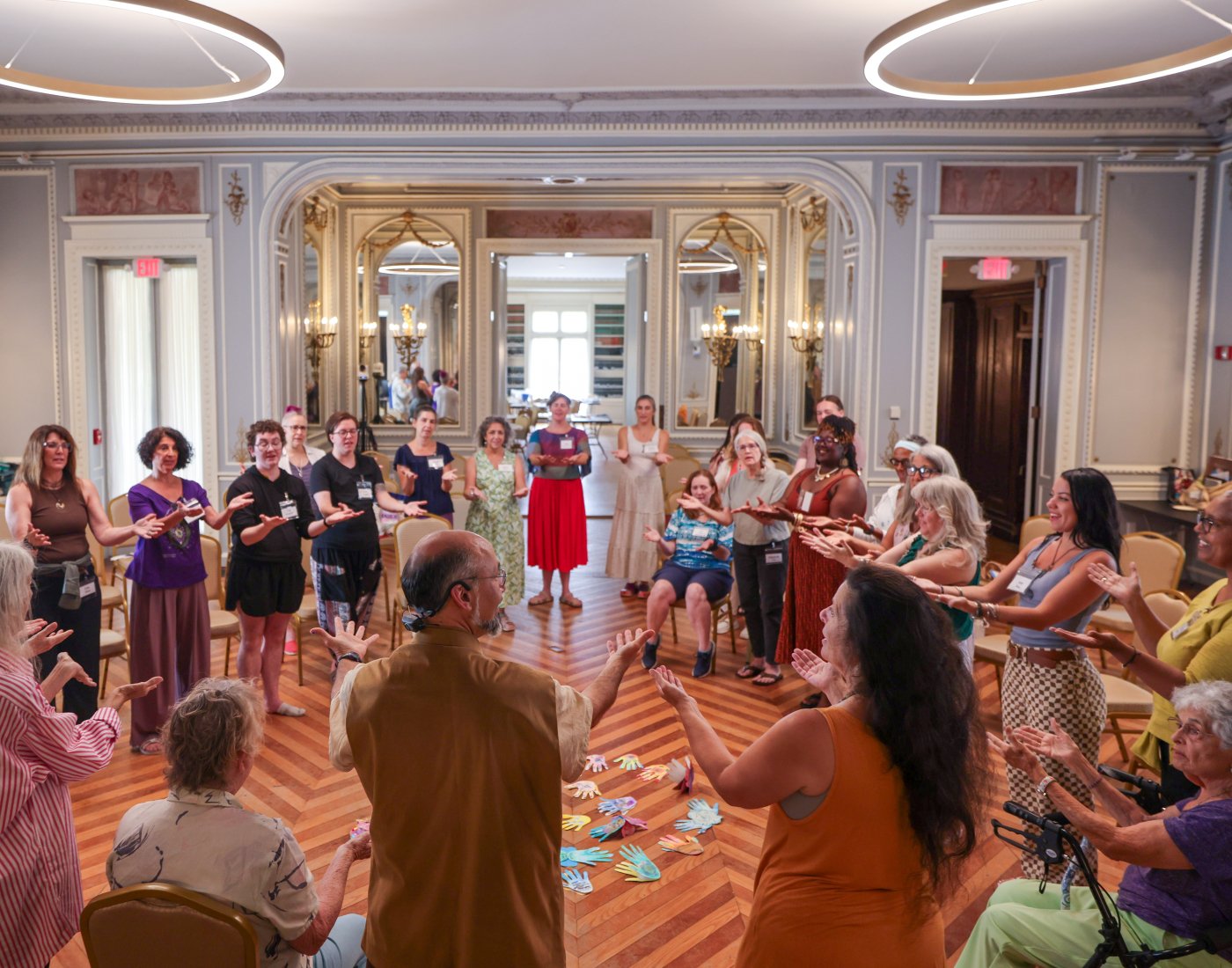 A large group of expressive arts students standing in a circle singing and with their arms extended outwards and palms up.