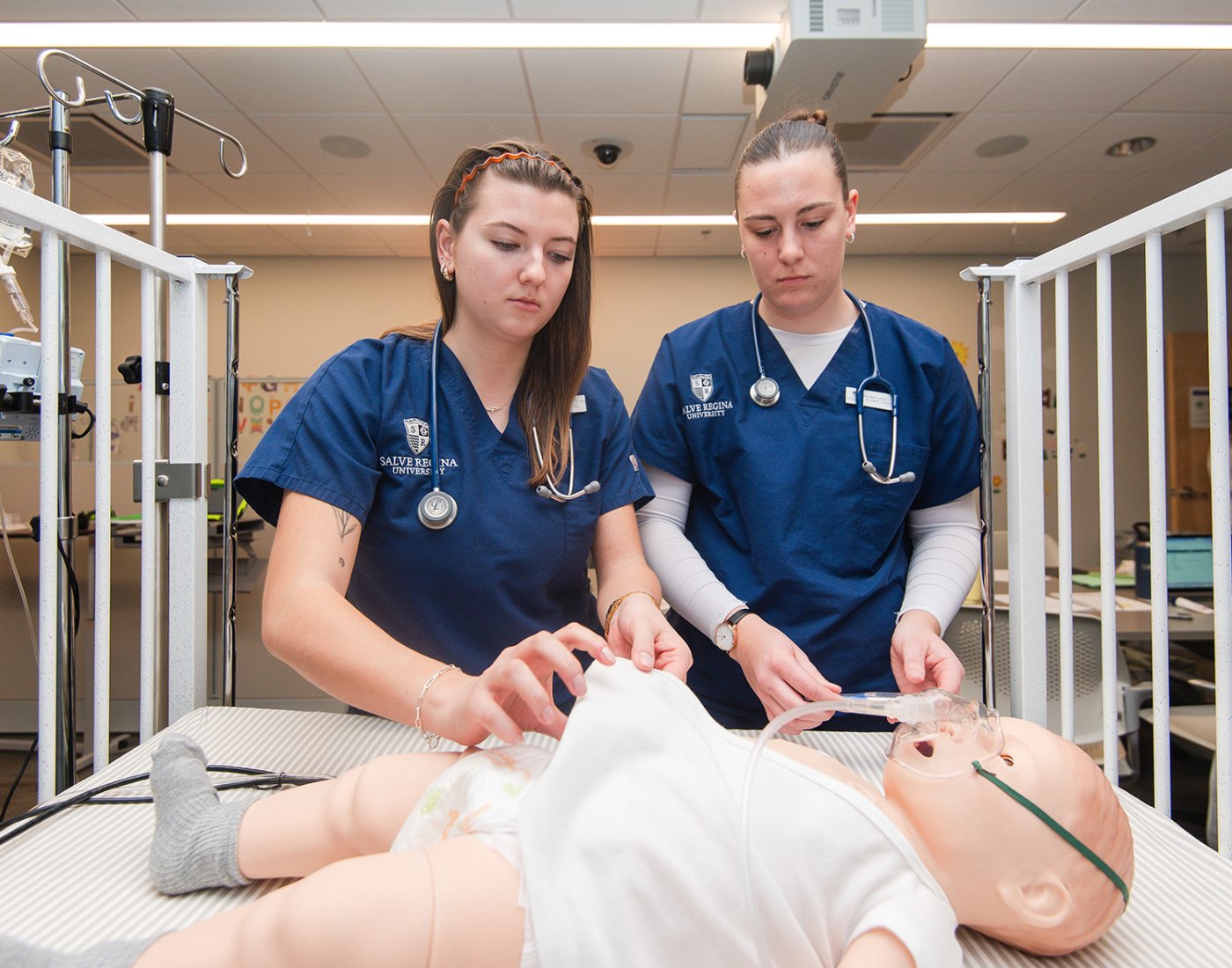Two nursing students in blue scrubs practice patient care on a newborn manikin in a simulated hospital room.