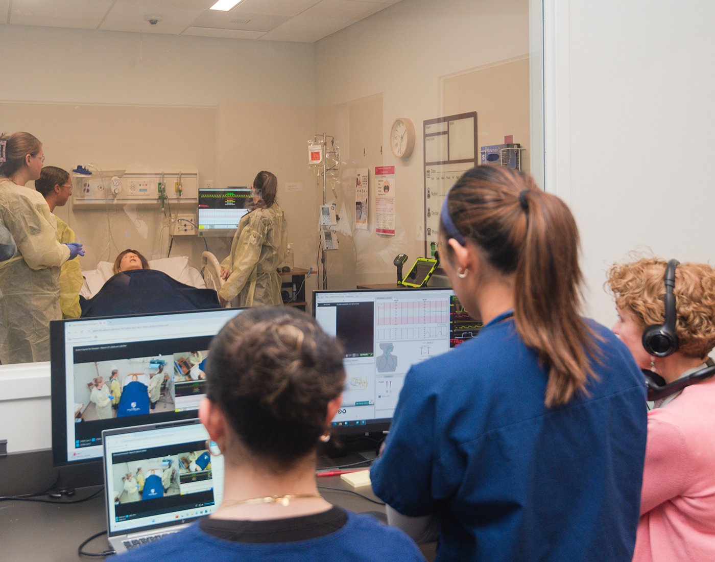 Nursing students observe a simulated patient scenario through a viewing window while instructors monitor the session from a debriefing room.