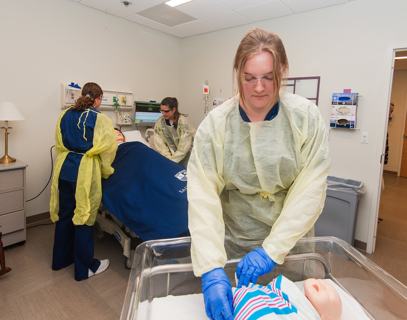 A student in a protective gown cares for a newborn manikin, while other students assist a female manikin on a hospital bed in the background.