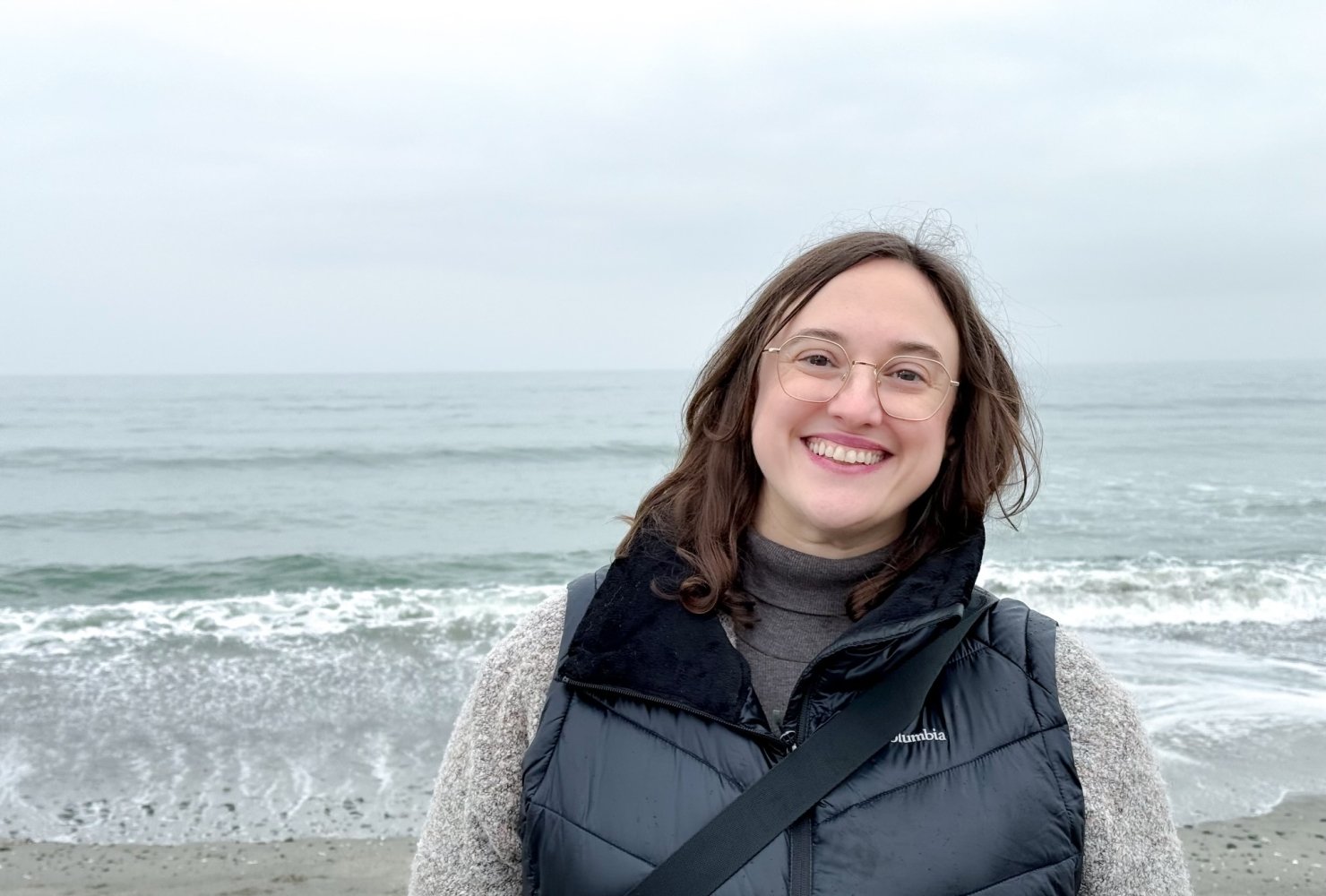 Photo of Brennan at the beach, smiling in a turtleneck and vest.