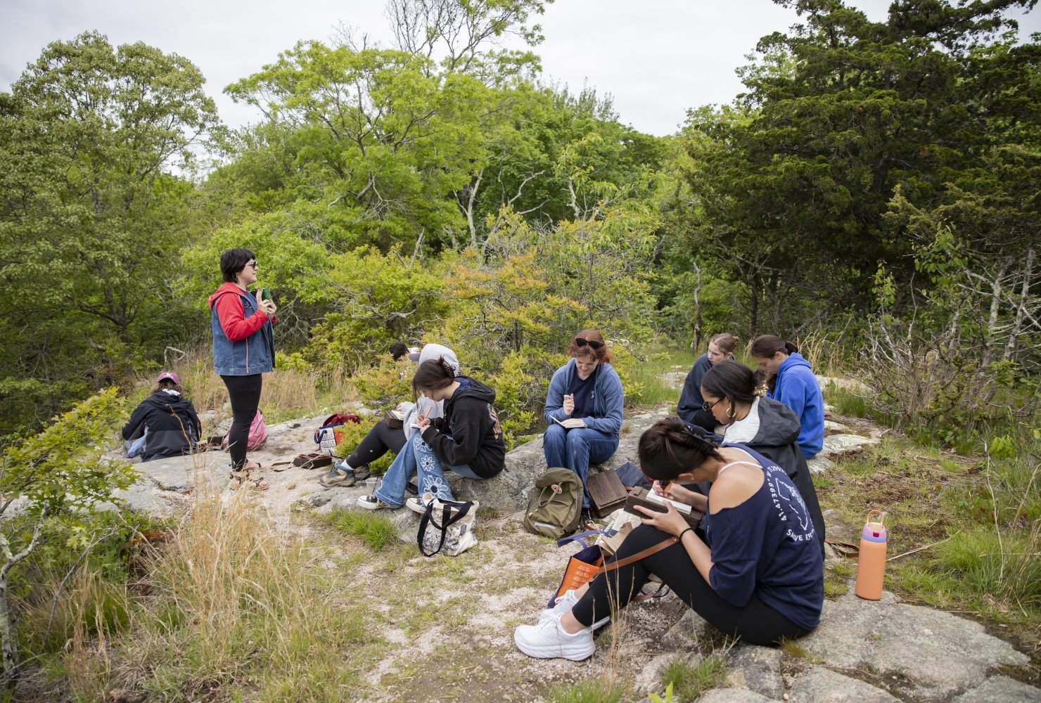 Group of students sitting in a green wooded area, drawing in their sketchbooks.