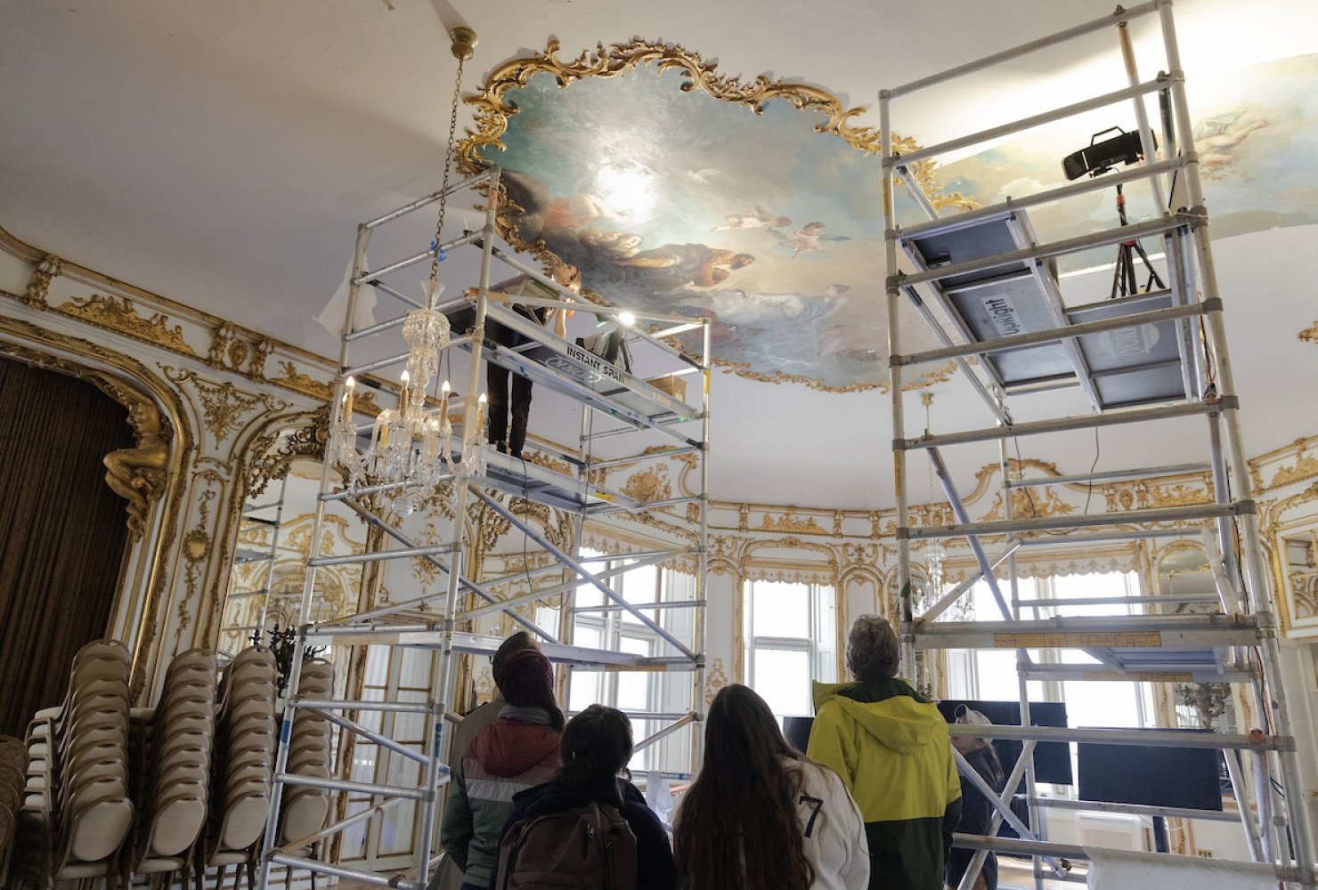 A group of people with their backs turned to the camera looking up at an oval-shaped ceiling painting in Ochre Court's ballroom.