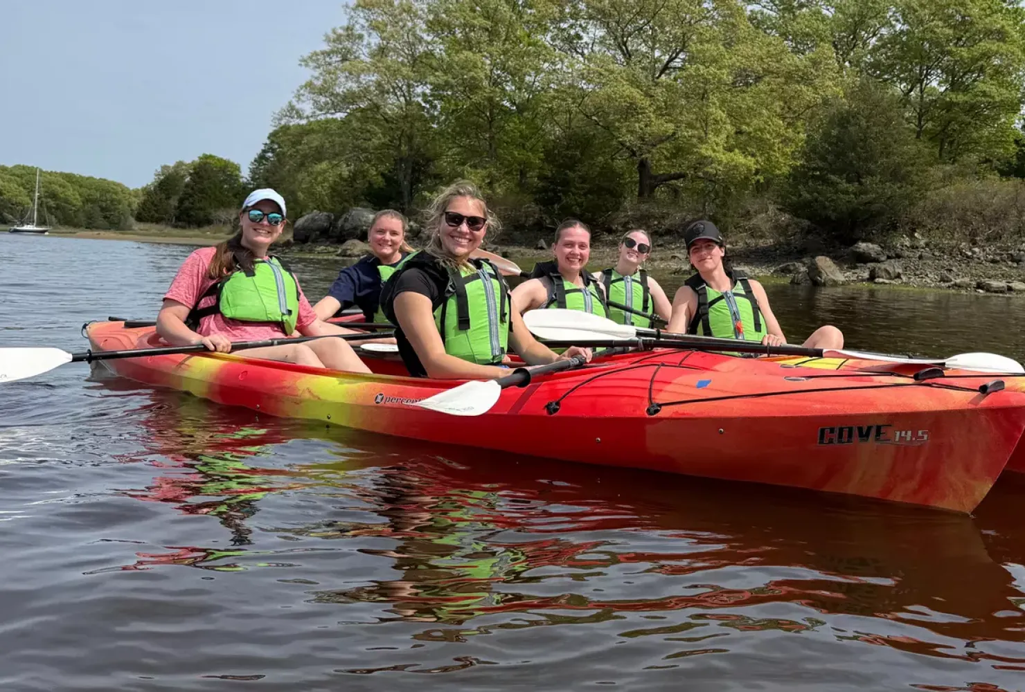 Group of six female students in lime green life vests sitting it two red kayaks on a lake with trees in the background, smiling at camera.