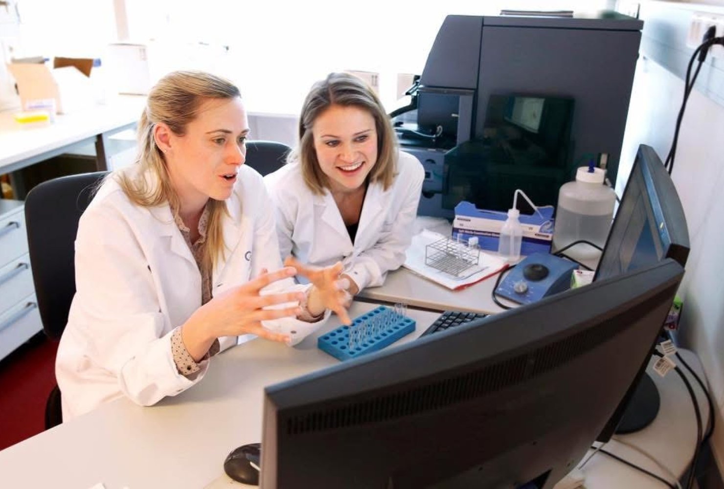 Jillian Wise and another collegue sitting in white lab coats discussing something they are viewing on the computer in front of them.