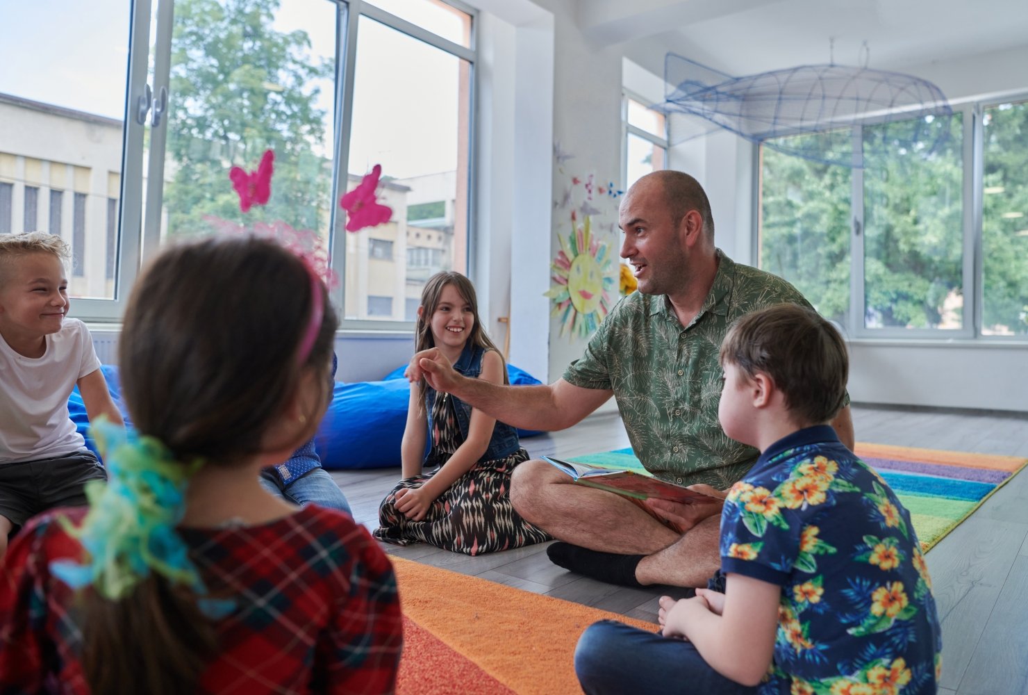 Male teacher teaching a group of young children, all sitting on the floor in a brightly lit classroom.