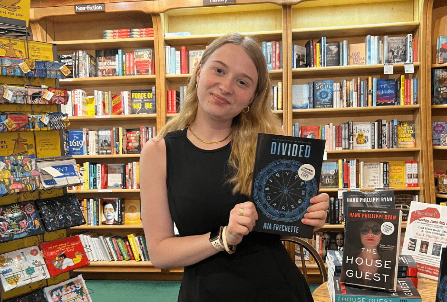 Ava wearing a black dress in a bookstore holding a copy of her first book "Divided"