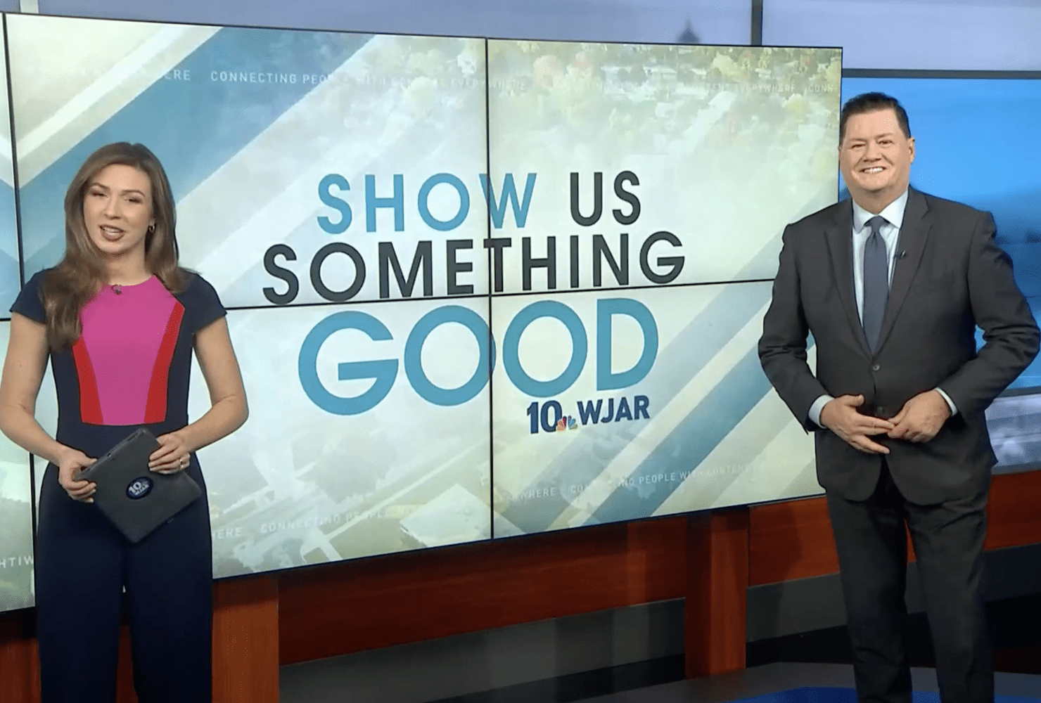 two news anchors standing in front of large tv on set that says 'Show us something good'