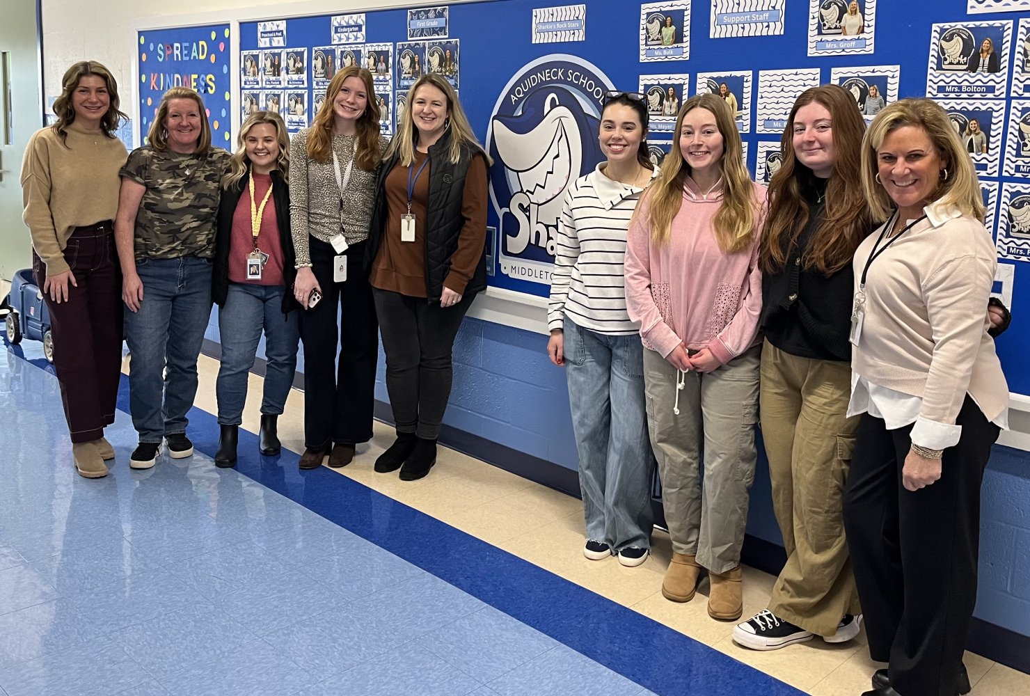 Nine education alums standing next to eachother smiling in front of blue bulletin board at Aquidneck Elementary School.