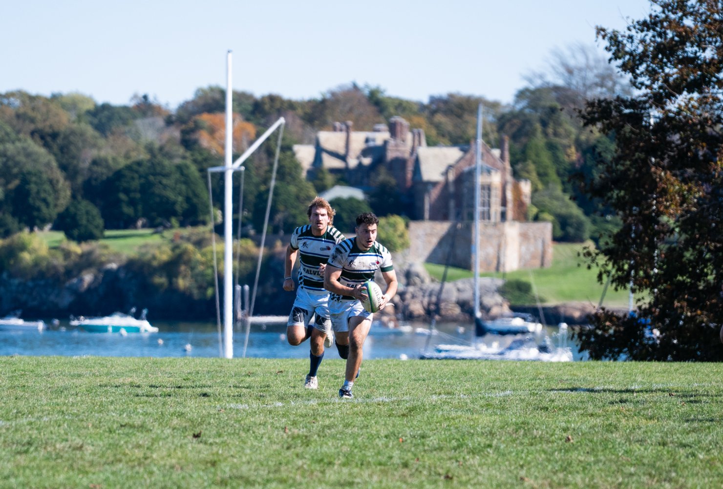 two men's rugby players on the field running after ball with ocean in background.