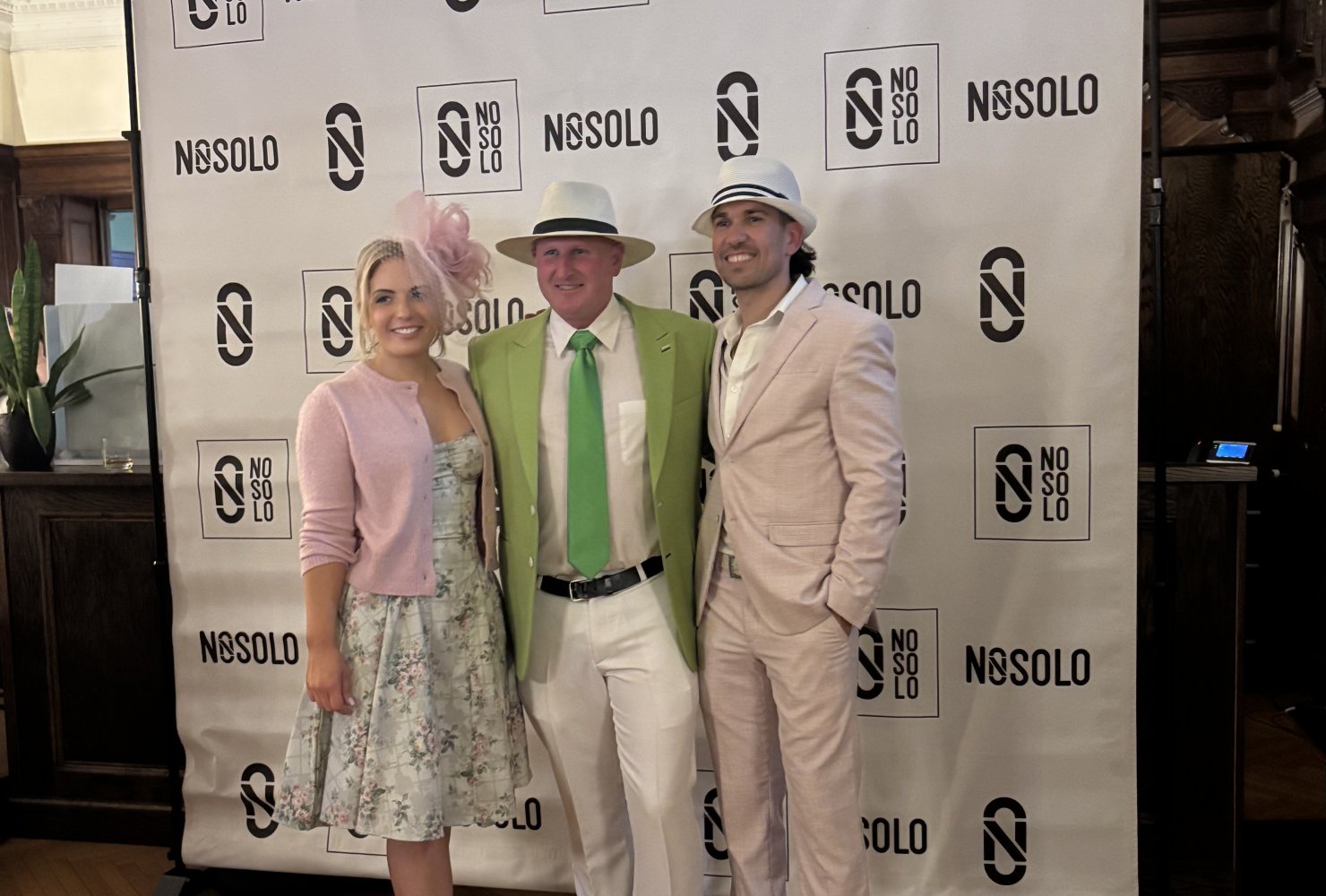 group photo of three people in pink and green outfits in front of a NOSOLO backdrop, for a Kentucky Derby fundraising event.