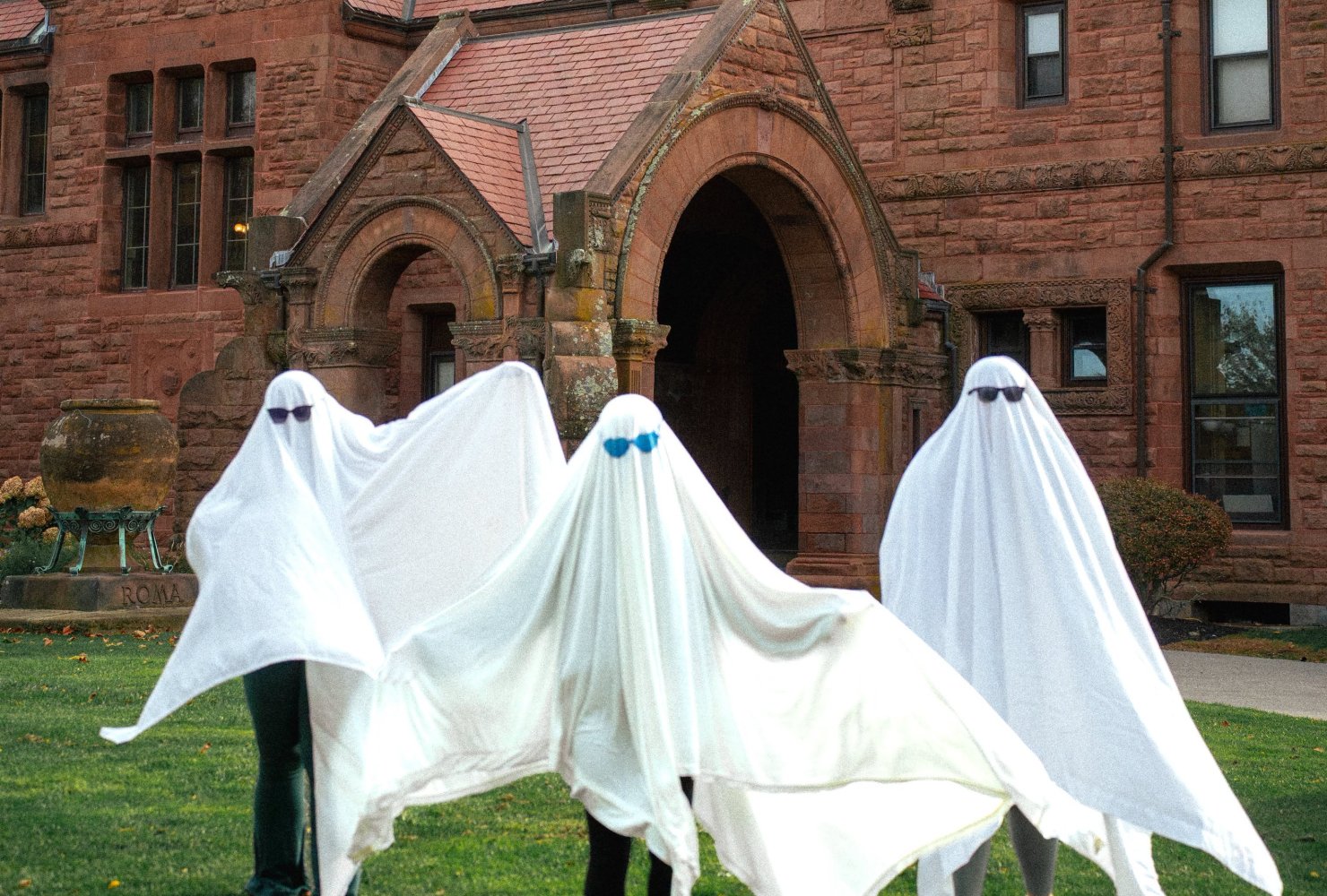 Three students dressed as ghosts in front of red building.