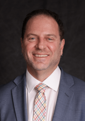 Professional headshot of Lorenz smiling in grey suit and colorful plaid tie.