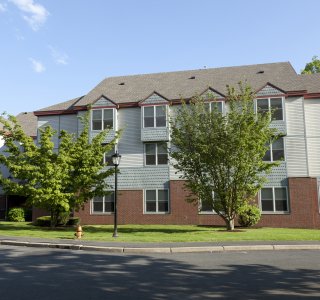 Three-story residential building with light siding, brick lower walls and trees in front.