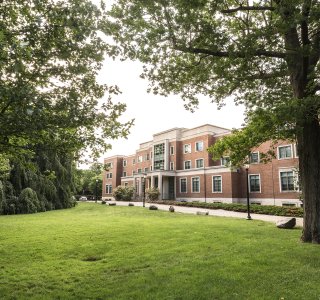 A brick building with large windows and a columned entrance, surrounded by a green lawn and tall leafy trees.