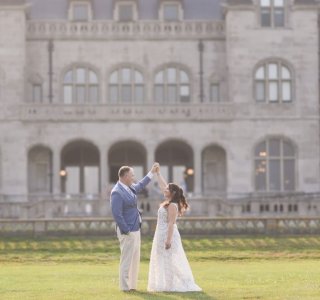 A couple in wedding attire stands on a lawn in front of a large historic stone mansion.