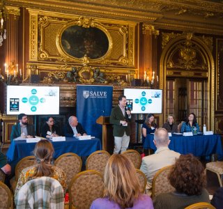 Panel discussion at a conference, with a speaker standing at a podium and seated panelists facing an audience.