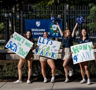 Four students stand outside a campus fence holding handmade signs in front of a banner that says "Welcome Seahawks."
