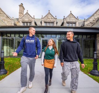 Three college students walk toward the camera outside a stone campus building on a sunny fall day.