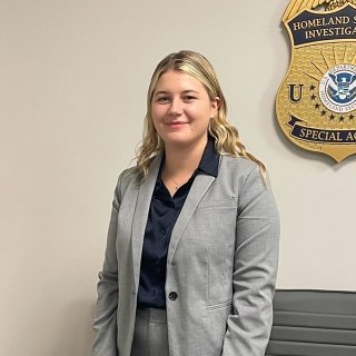 Photo of Lia in a gray suit standing in front of an american flag and a Homeland Securit Investigations Special Agent plaque