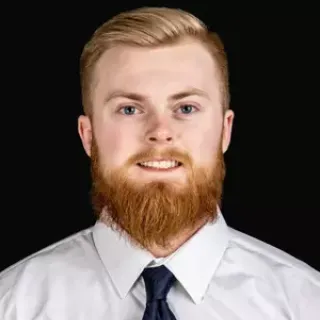 Male student with reddish hair and beard, in white shirt and dark solid color tie looking at camera