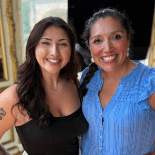 Acevedo (left) in a black top and Jennifer De Leon in a blue dress together and smiling at the camera.