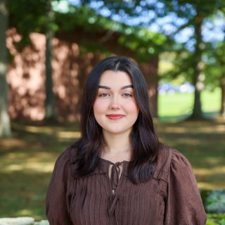 Young alumna, brown hair, brown shirt, standing outside smiling