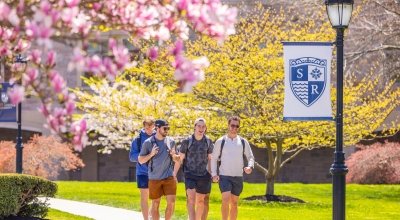 Four mail students walking on campus by tree in full pink bloom, near light with salve crest banner