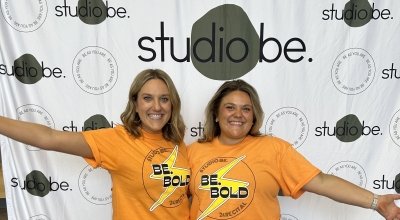 Two girls smiling in matching orange t-shirts in front of a Studio Be backdrop in black and white.