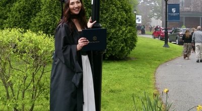 Acevedo wearing her full regalia, holding her diploma smiling next to a lamppost with Salve's logo/flag