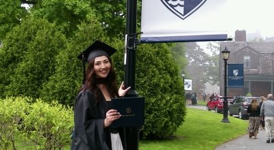 Acevedo wearing her full regalia, holding her diploma smiling next to a lamppost with Salve's logo/flag