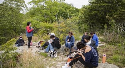 Group of students sitting in a green wooded area, drawing in their sketchbooks.