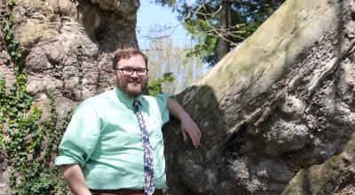 Ben Hohman in an aqua colored button down shirt and pink and blue tie, smiling with his arm rested up against a tree.