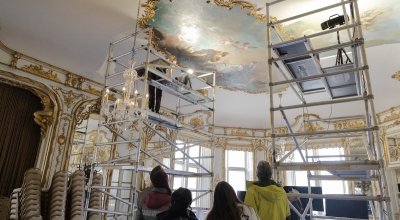 A group of people with their backs turned to the camera looking up at an oval-shaped ceiling painting in Ochre Court's ballroom.