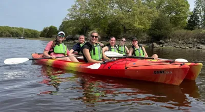 Group of six female students in lime green life vests sitting it two red kayaks on a lake with trees in the background, smiling at camera.