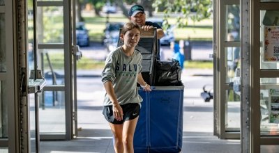 Student in Salve shirt pulling a cart of her stuff behind her as she walks into her residence hall.