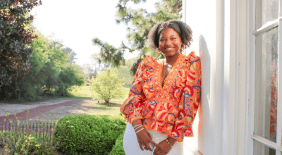 Professional landscape shot of Elodie in colorful orange blouse, smiling while leaning against a white building with trees in background.