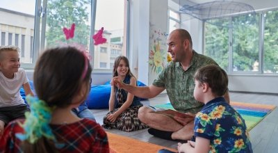 Male teacher teaching a group of young children, all sitting on the floor in a brightly lit classroom.