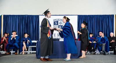 Dever receiving his diploma from President Armstrong in full regalia, with blue drapery and a Salve sign in the background.