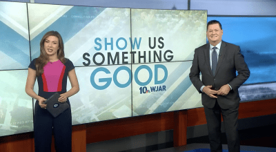 two news anchors standing in front of large tv on set that says 'Show us something good'