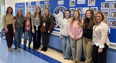 Nine education alums standing next to eachother smiling in front of blue bulletin board at Aquidneck Elementary School.