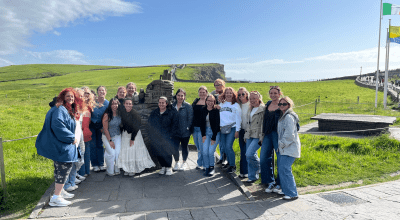 female students and faculty smile for a group photo in Ireland in front of a field of grass.
