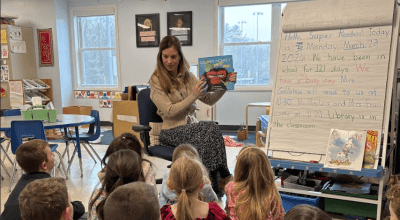 Teacher reads a picture book to students seated on a classroom rug.